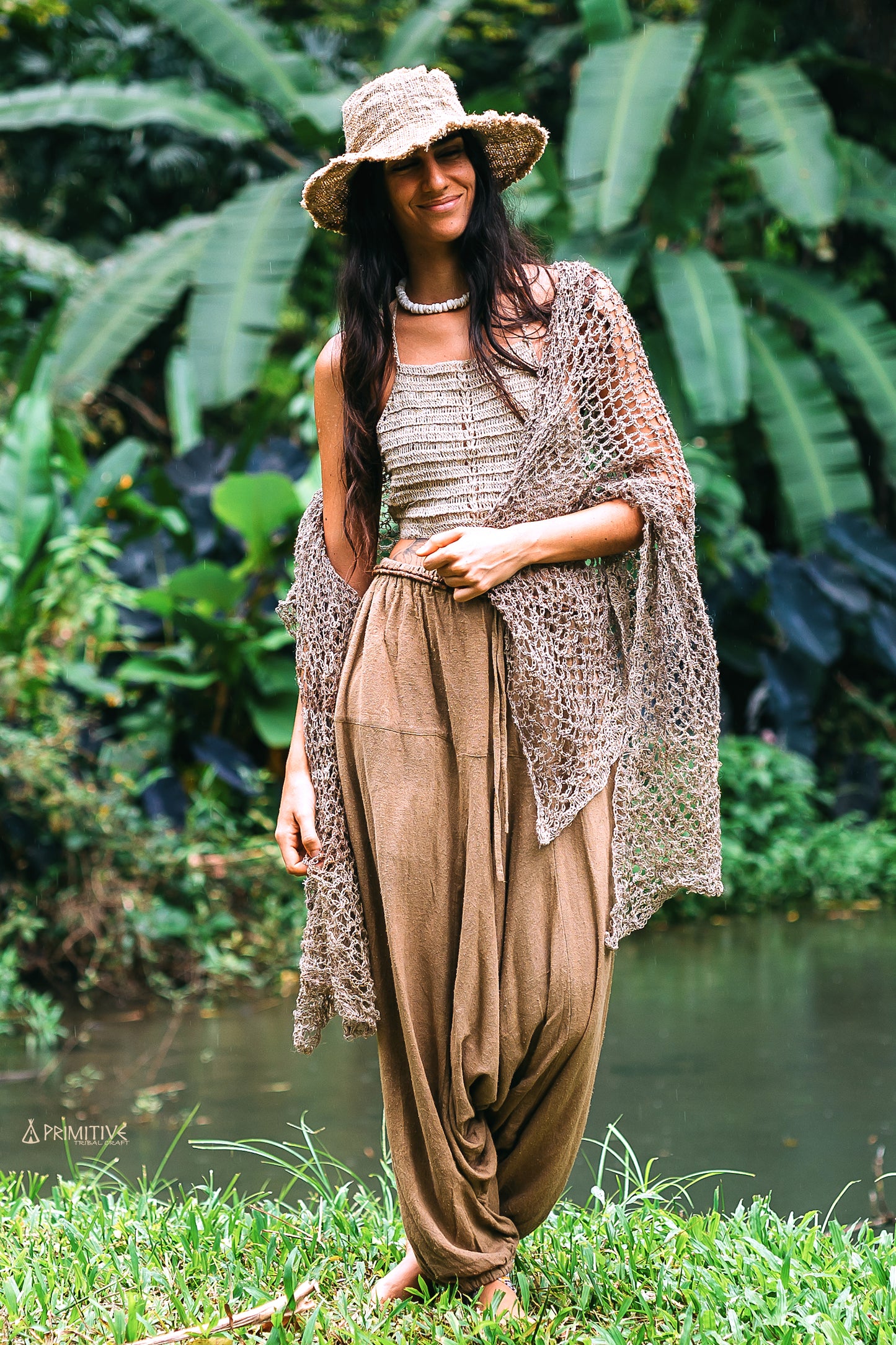 A women standing in a lush green environment wearing a large knitted nettle shawl in a natural color, and raw silk afghani pants