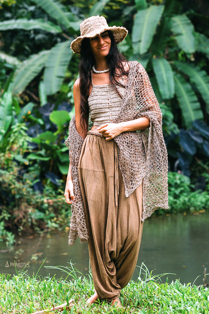 A women standing in a lush green environment wearing a large knitted nettle shawl in a natural color, and raw silk afghani pants