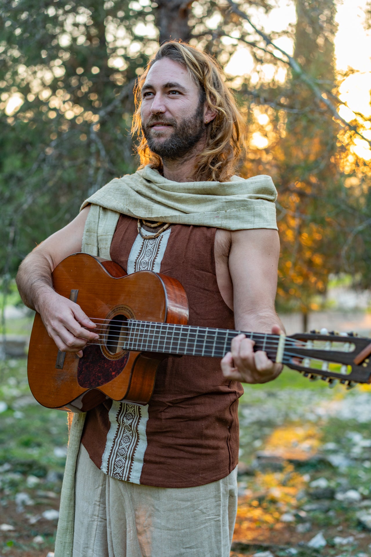 hippie Man wearing a handwoven raw silk tank top with a vertical tribal block print, holding an acoustic guitar outdoors at sunset.