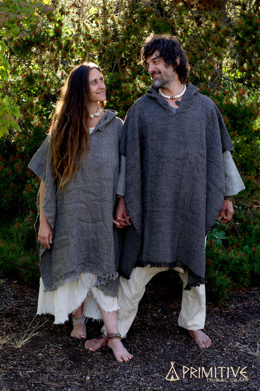 Two people wearing woven wool poncho with hood, standing outdoors with greenery in the background.
