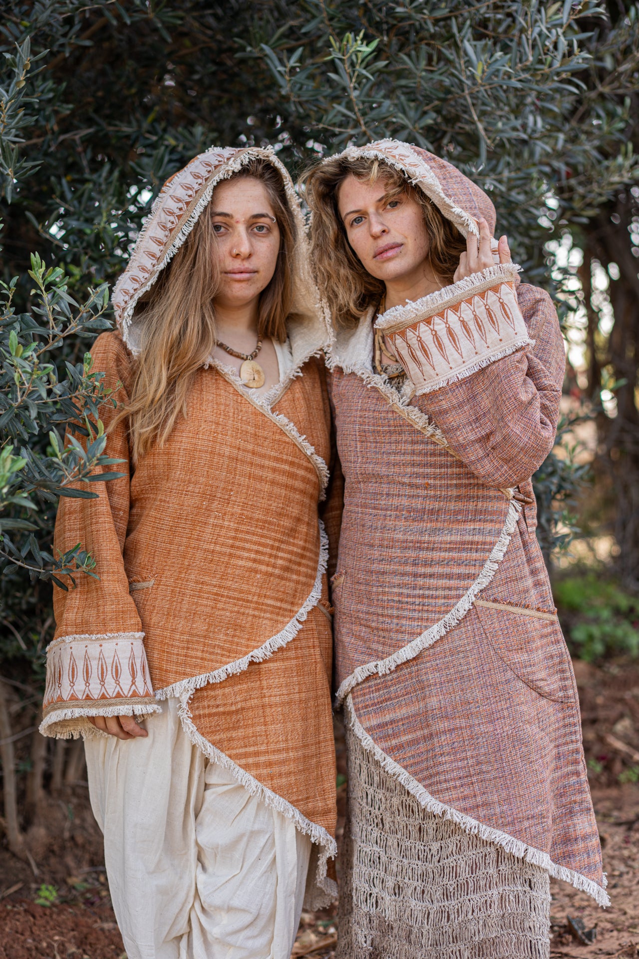 Two women wearing earth-tone hooded Khadi cotton jackets with rustic fringe and tribal embroidery details in a natural setting