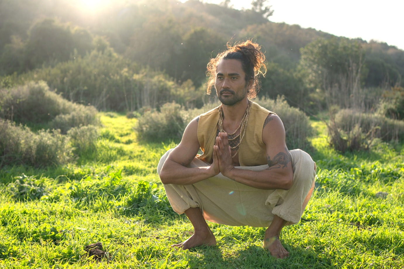 Man wearing camel khadi cotton vest and loose natural pants, squatting outdoors in a prayer pose at golden hour.