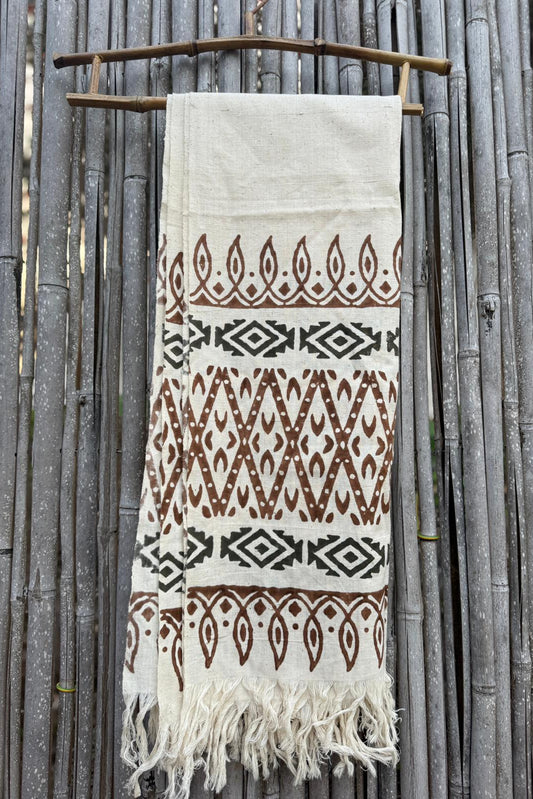 Handwoven khadi cotton shawl in natural cream with brown and black tribal prints and fringed ends, hanging on a bamboo fence.