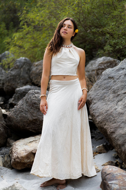 Woman standing on river rocks wearing a white handloom khadi cotton maxi wrap skirt and matching halter crop top. Earthy bohemian summer fashion.