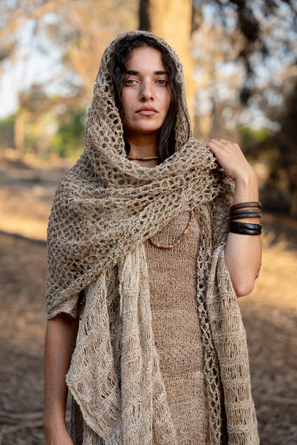A woman wearing a large, open-knit shawl made from raw Wild Himalayan Nettle fiber, draped over her head as a hood in a natural outdoor setting.