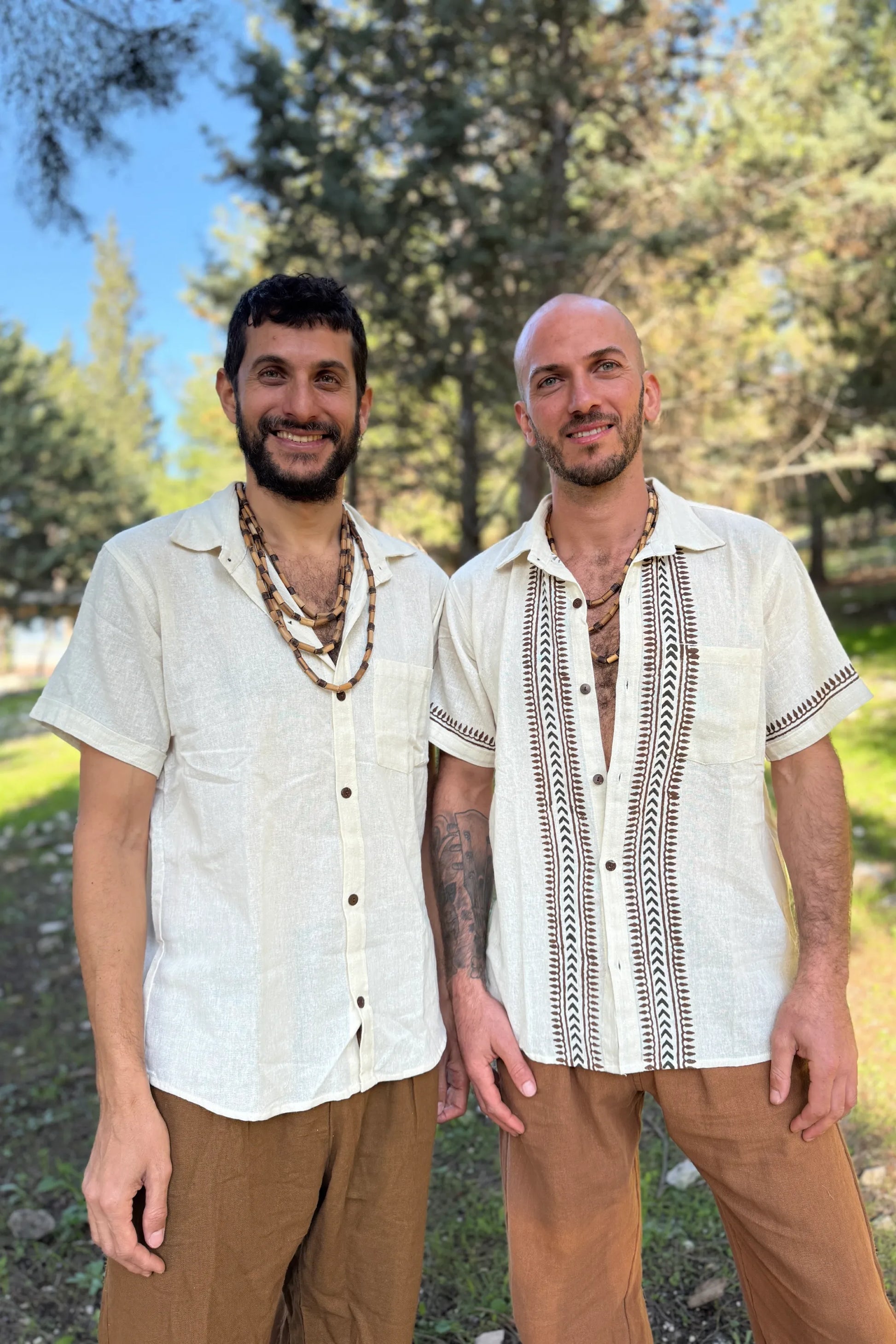 Two men standing outdoors wearing white short-sleeve cotton button-down shirts, one plain and one with hand-stamped tribal block prints.
