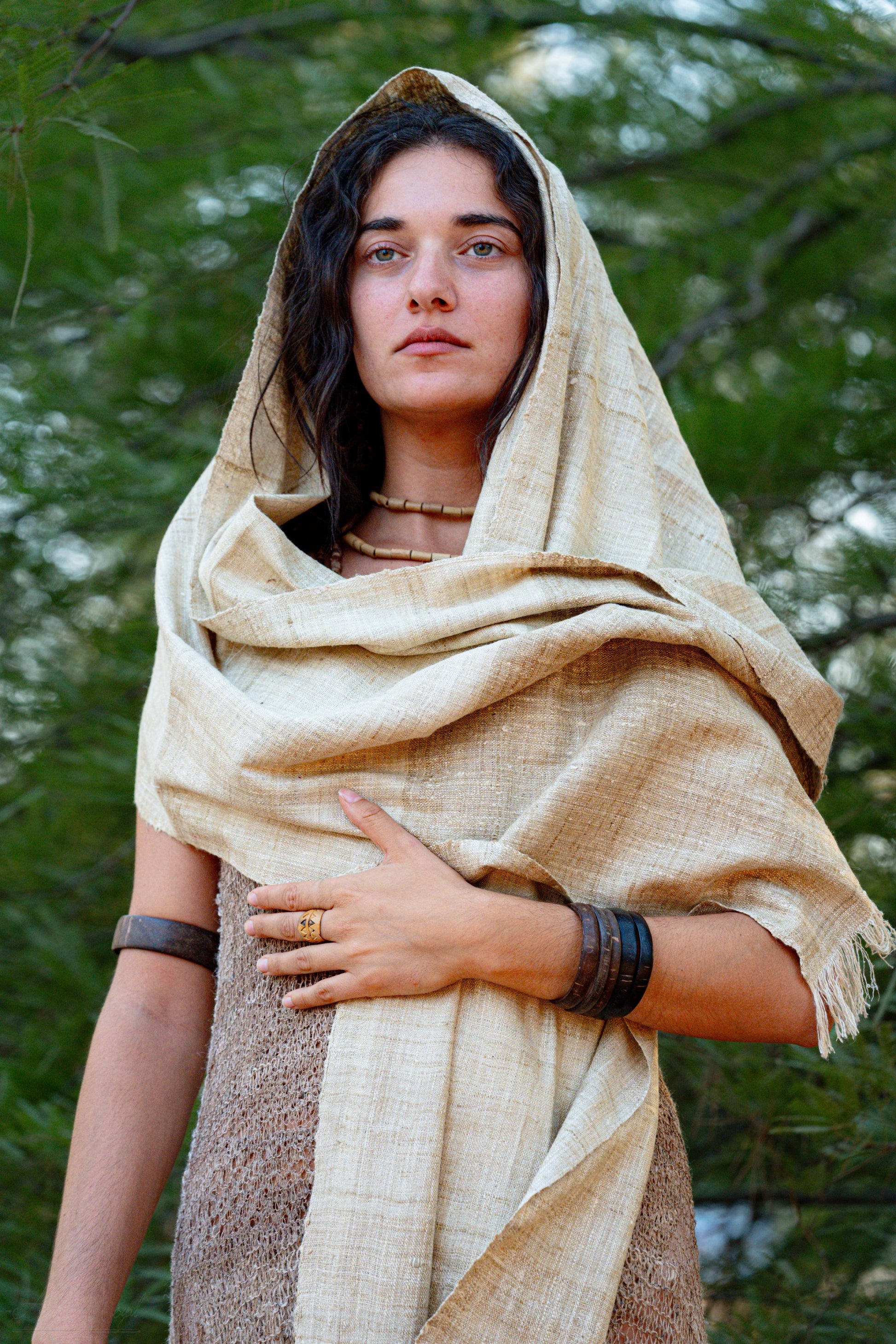 Boho Woman wearing a natural handwoven mulberry raw silk shawl styled as a hood, photographed outdoors among green trees.