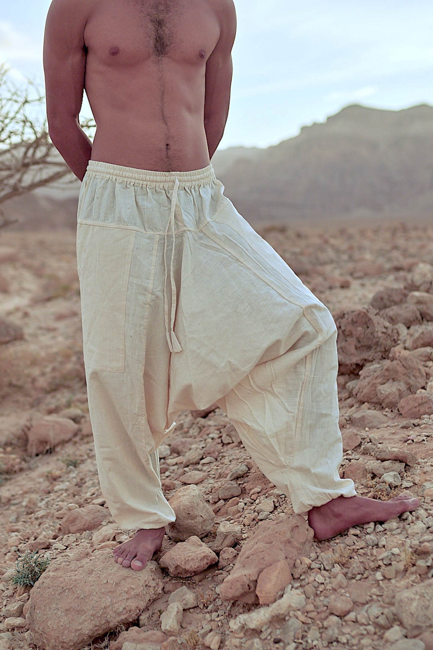 Unisex natural cotton harem pants in neutral color with drop-crotch fit, drawstring waist and relaxed legs, photographed outdoors in a desert setting.