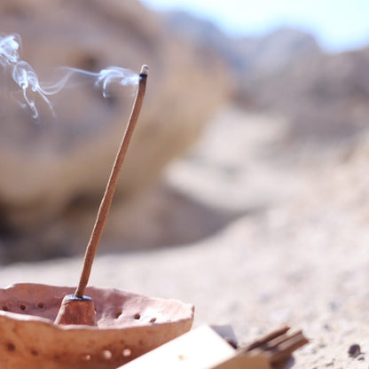 Natural incense stick burning in a ceramic copaler with a majestic desert landscape background, emitting soft white smoke.