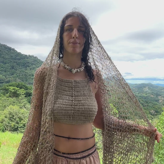 A woman wrapped in a large, beige, open-knit shawl made of wild nettle, standing in a sun-drenched forest, looking directly at the camera.