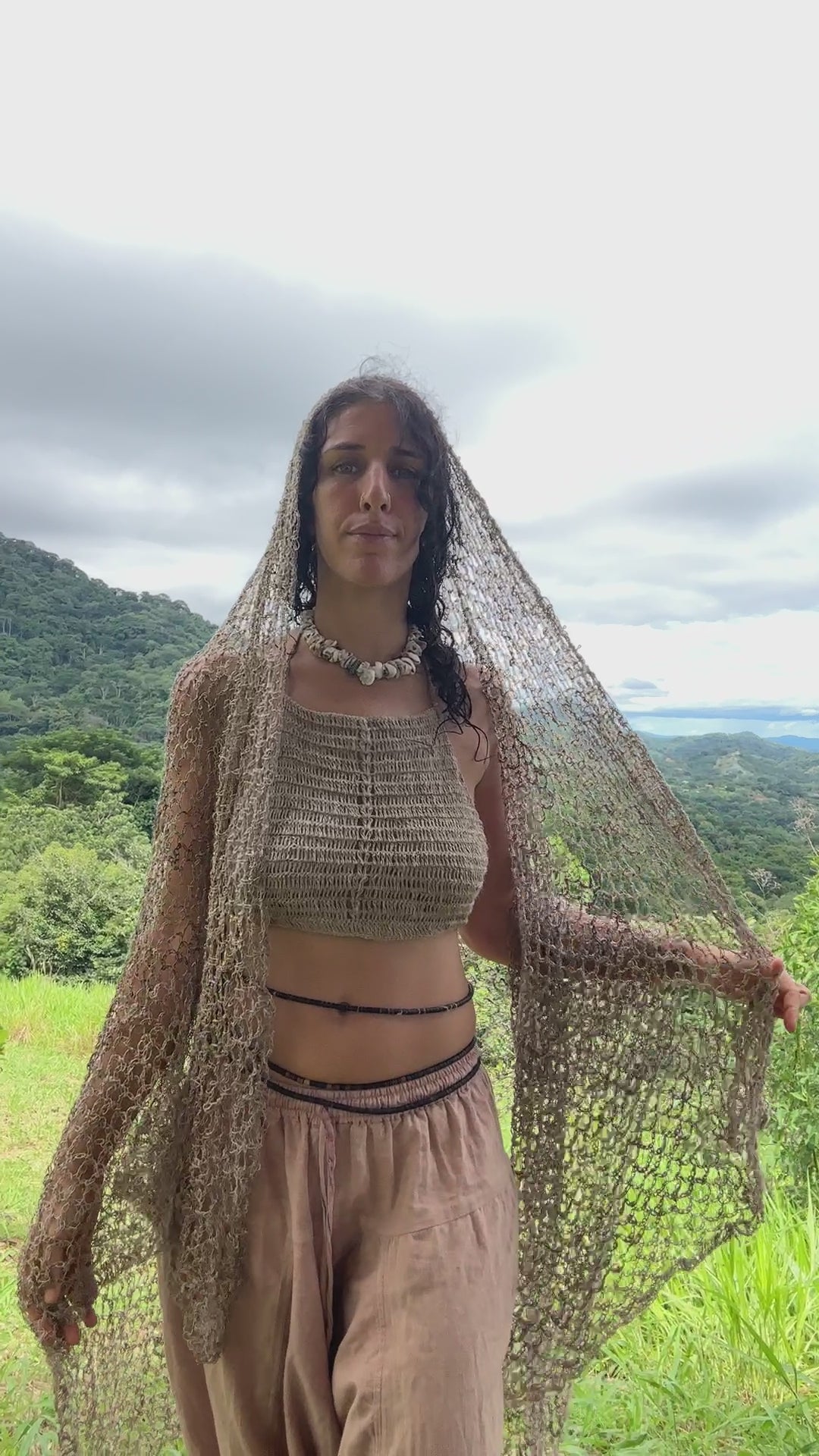 A woman wrapped in a large, beige, open-knit shawl made of wild nettle, standing in a sun-drenched forest, looking directly at the camera.