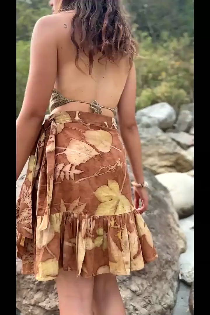 A short video showing a woman turning gracefully on river rocks, modeling a handmade botanical print wrap skirt with natural leaf designs and a ruffled hem.