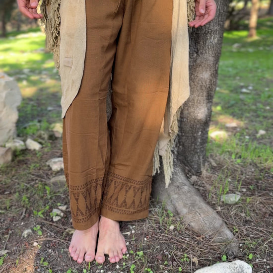 Detailed view of earth-toned brown cotton pants for men, featuring tribal tipi patterns on the leg cuffs, styled in a natural outdoor setting