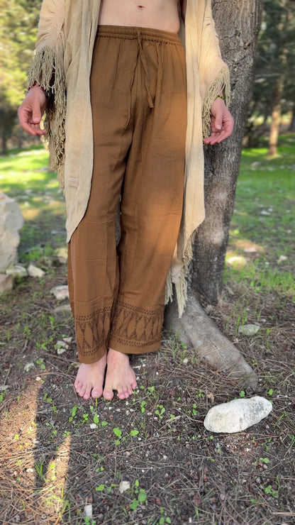Detailed view of earth-toned brown cotton pants for men, featuring tribal tipi patterns on the leg cuffs, styled in a natural outdoor setting