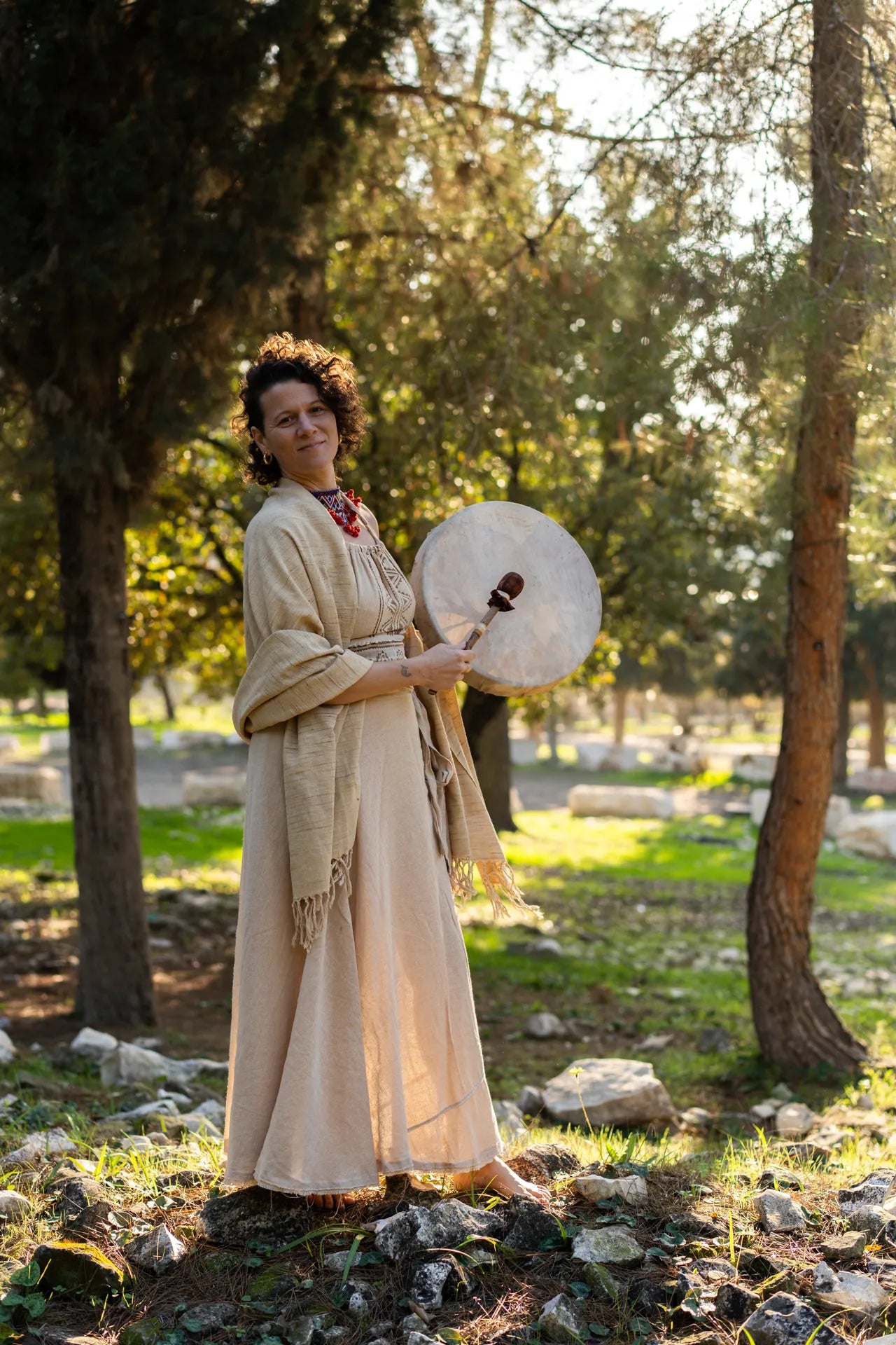 Woman wearing a natural boho halter top and wrap skirt with a fringed shawl, holding a ceremonial drum outdoors.