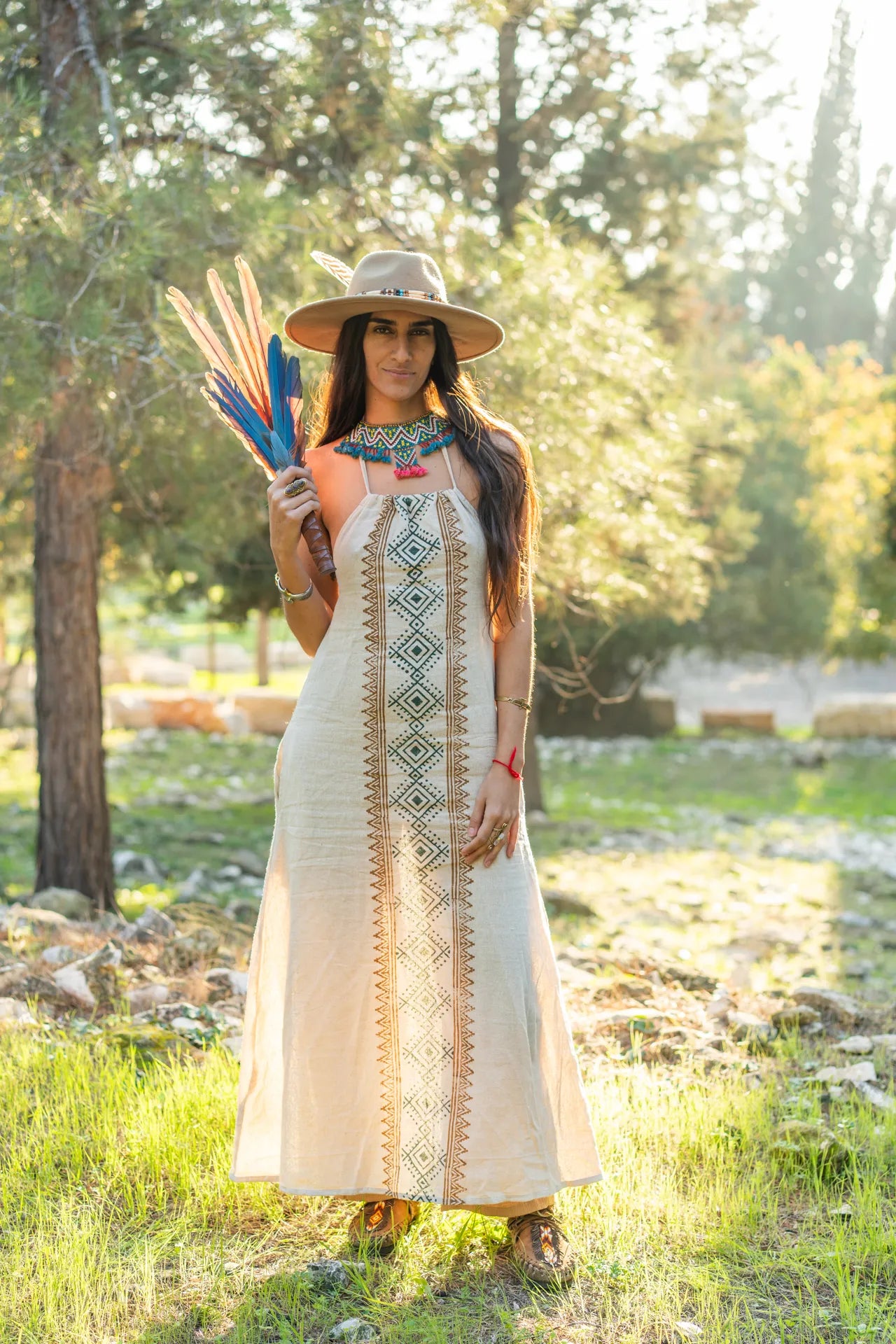 A striking natural cotton dress featuring a vertical tribal block print design, styled with a bohemian hat and a feather fan in a sun-drenched forest.