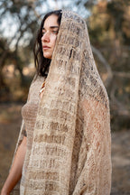 Close-up of a model wrapped in a thin natural nettle shawl with an airy open weave and striped texture, photographed in nature