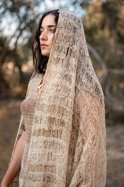 Close-up of a model wrapped in a thin natural nettle shawl with an airy open weave and striped texture, photographed in nature