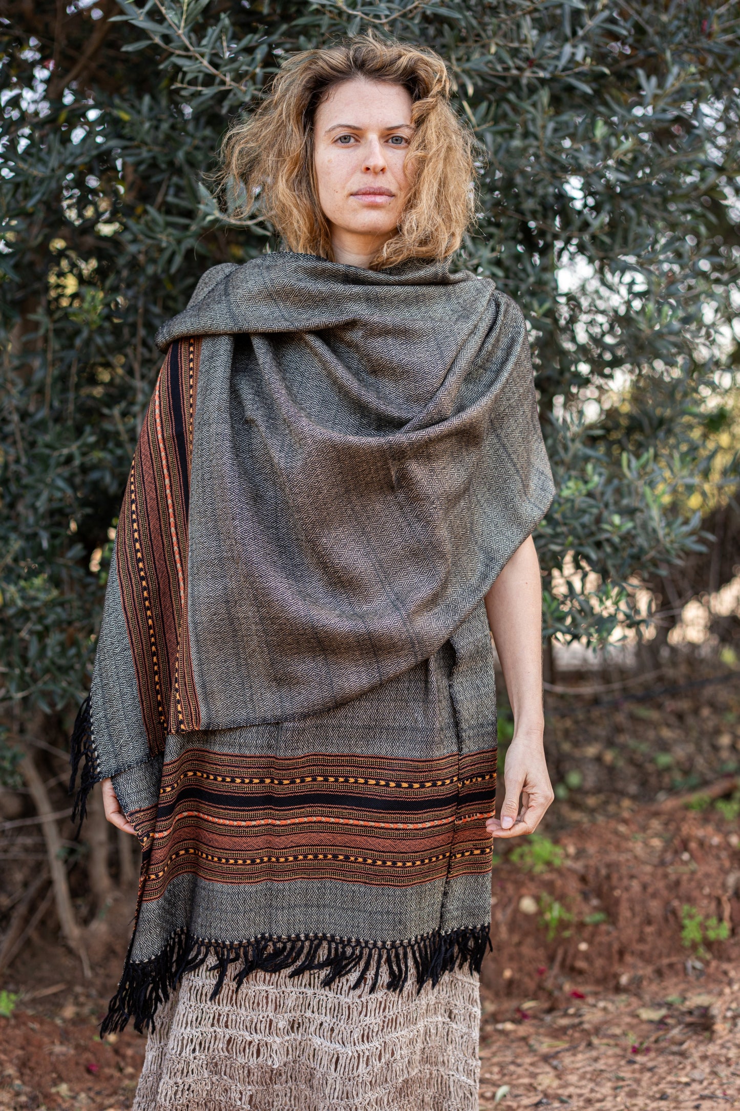Grey handwoven wool shawl featuring a detailed orange and black tribal border. Authentic Himalayan winter wrap with fringes styled on a woman in nature.