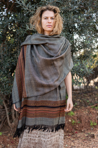 Grey handwoven wool shawl featuring a detailed orange and black tribal border. Authentic Himalayan winter wrap with fringes styled on a woman in nature.