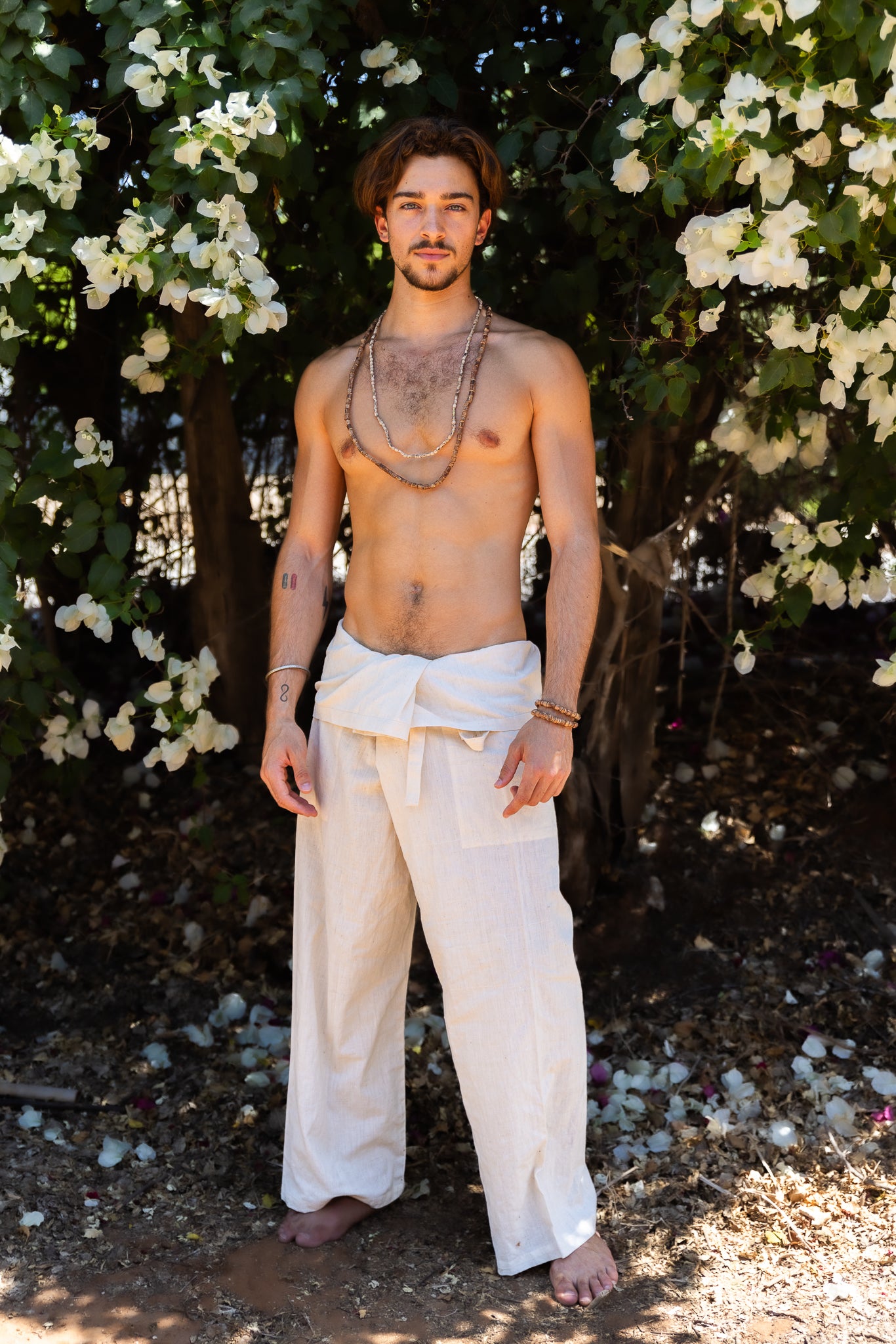 Man in white fisherman pants standing in a natural setting with flowers and trees.