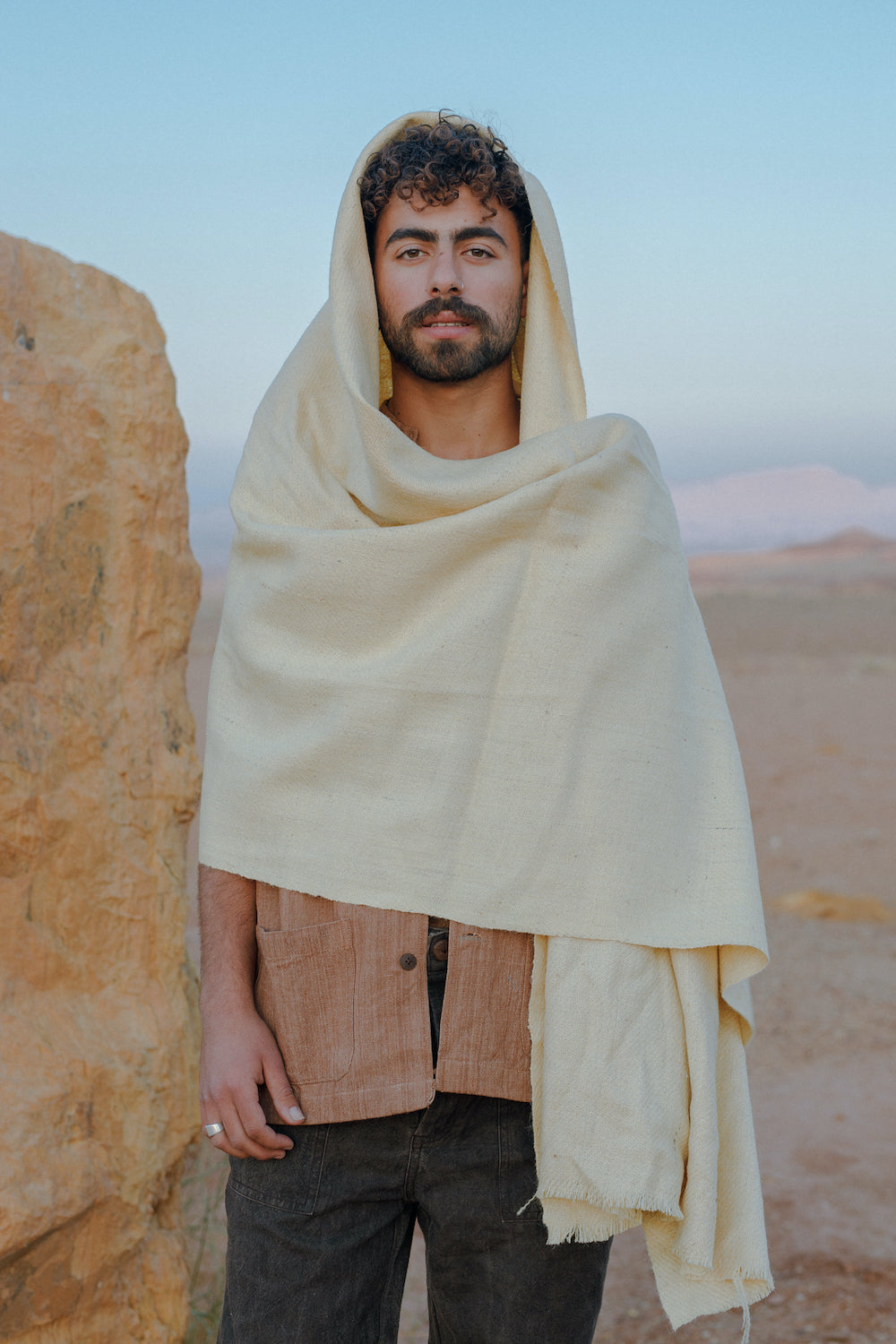 Man wearing a cream-colored Ancient Path Shawl ⫸ Handspun Handwoven Khadi Wool draped over his head and shoulders, standing outdoors near rocky terrain under a clear sky.