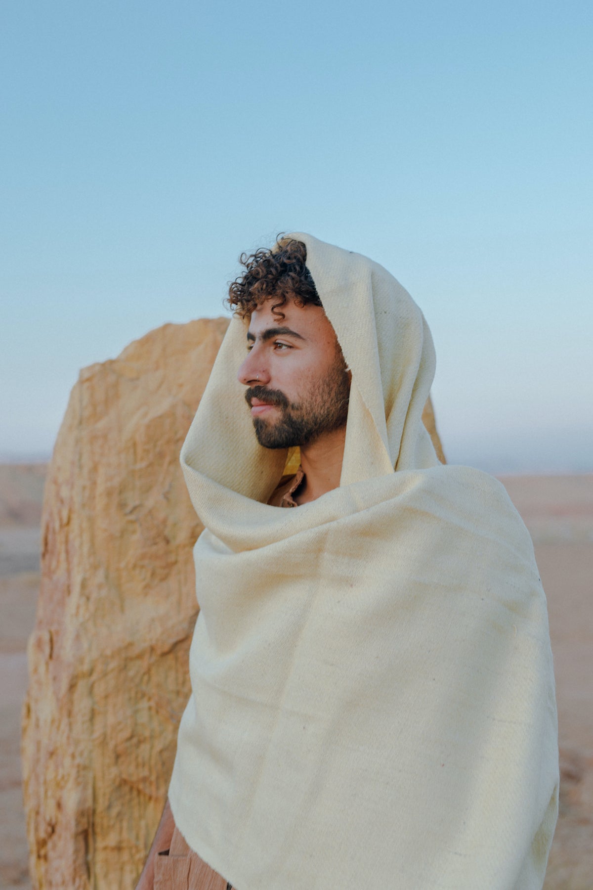 Man wearing a natural beige shawl draped over his head and shoulders, standing outdoors near a rock formation, showcasing the Ancient Path Shawl Handspun Handwoven Khadi Wool.