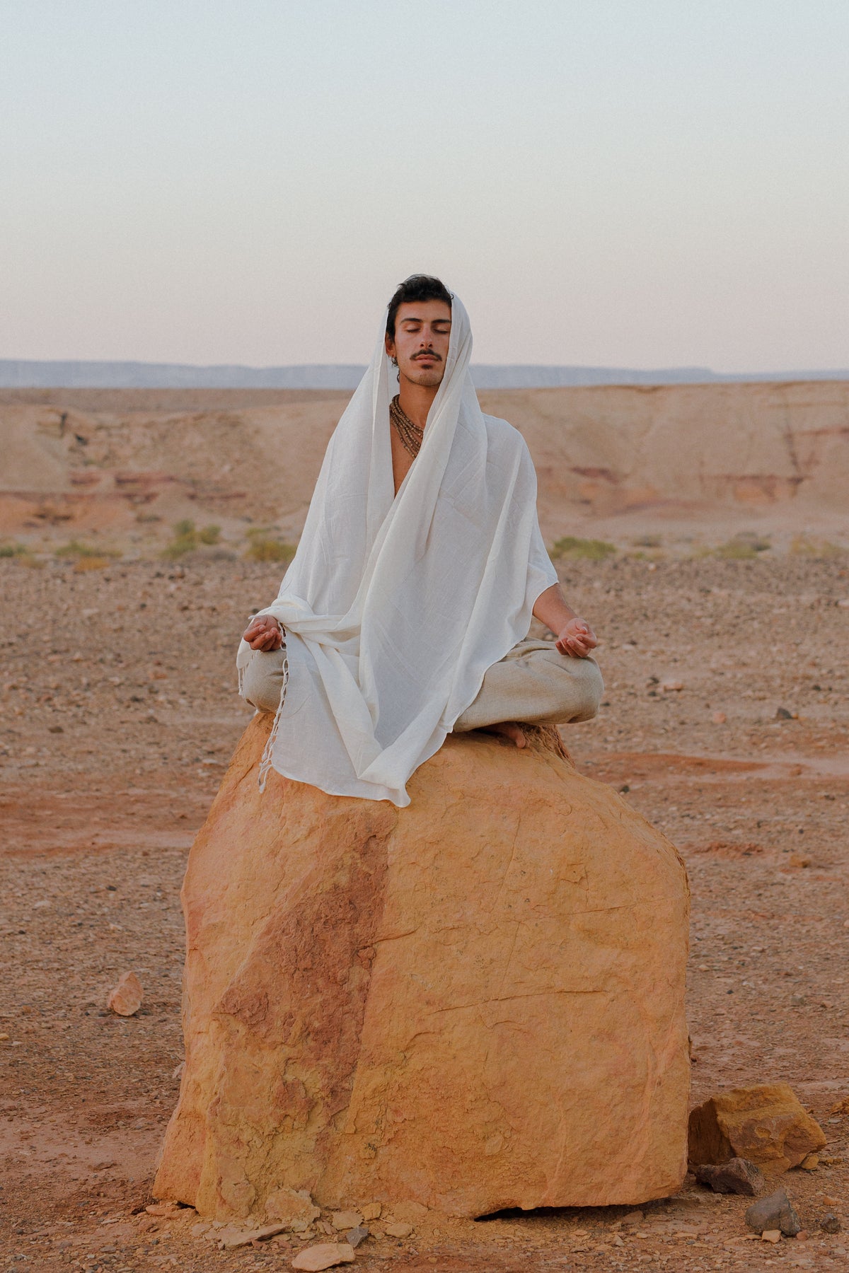 A person meditating outdoors wrapped in a Handwoven Cotton Shawl Traditional Weaving Hand Spun and Handwoven Cotton, sitting cross-legged on a large rock in a natural desert landscape.