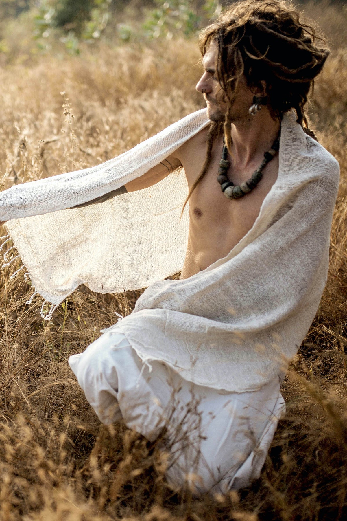 Man with dreadlocks wearing a necklace and wrapped in a natural woven cotton shawl ⋙⋗, seated outdoors in a dry grassy field during golden hour.