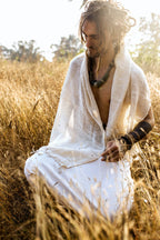 Man sitting in a sunlit field wearing a natural woven cotton shawl ⋙⋗ draped over his shoulders paired with white clothing and rustic jewelry