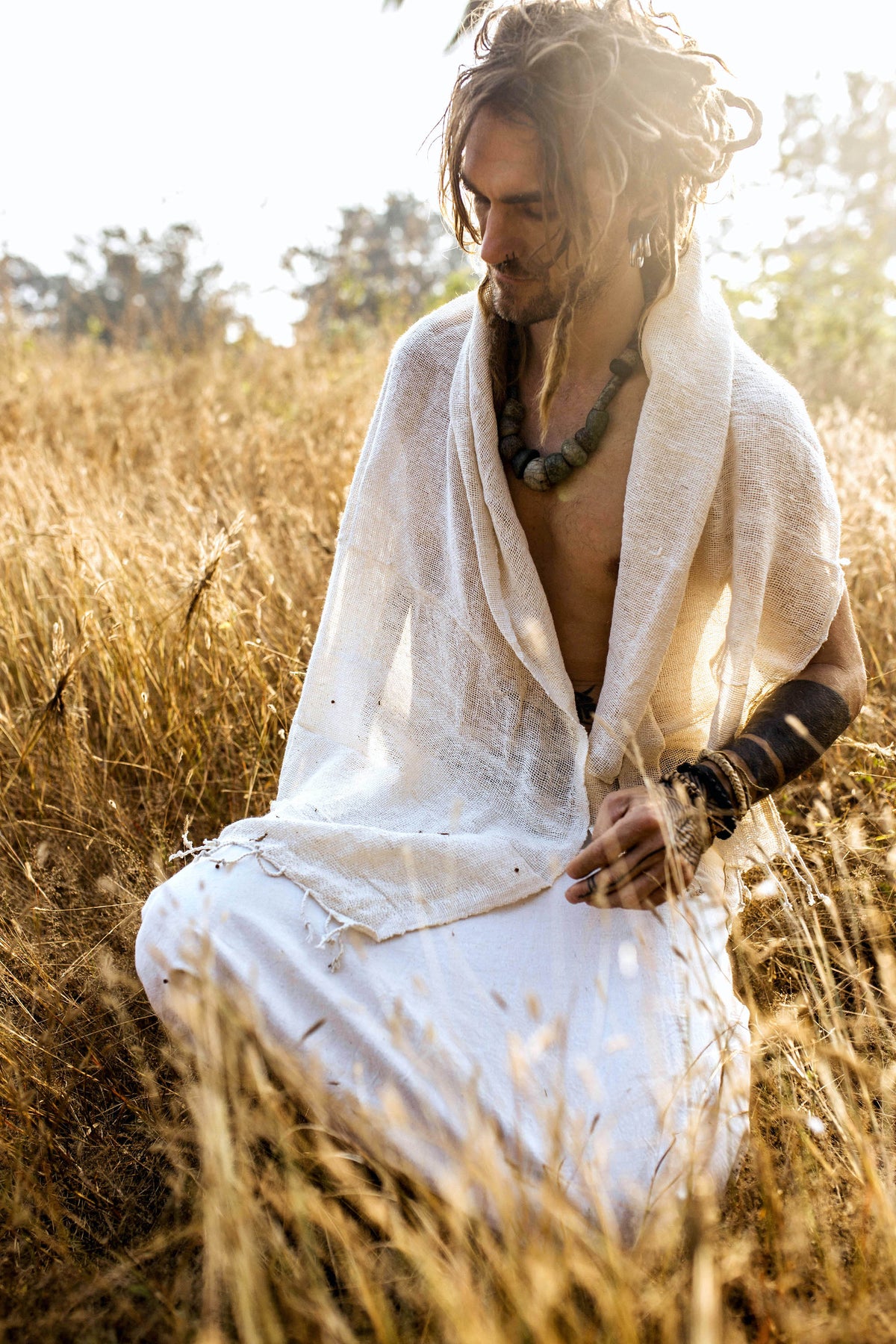 Man sitting in a sunlit field wearing a natural woven cotton shawl ⋙⋗ draped over his shoulders paired with white clothing and rustic jewelry