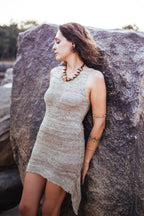 Model in sleeveless Himalayan nettle yarn dress, natural color knit with pointed side hem, leaning against rock outdoors.